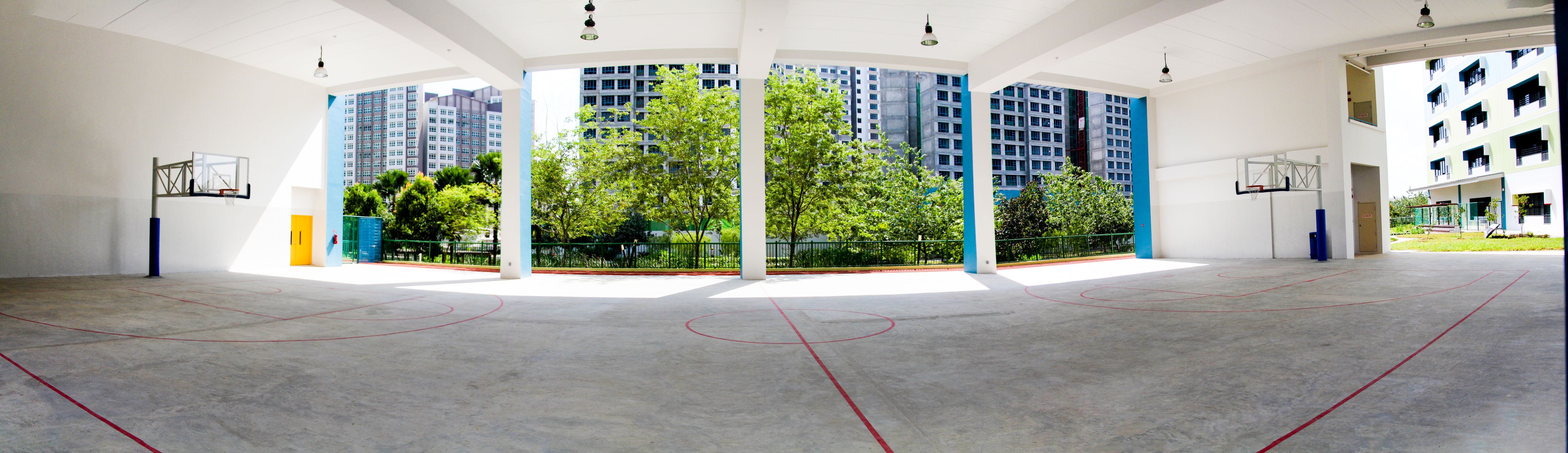 Panoramic view of an empty covered basketball court with two hoops, red court lines, and tall buildings visible outside.
